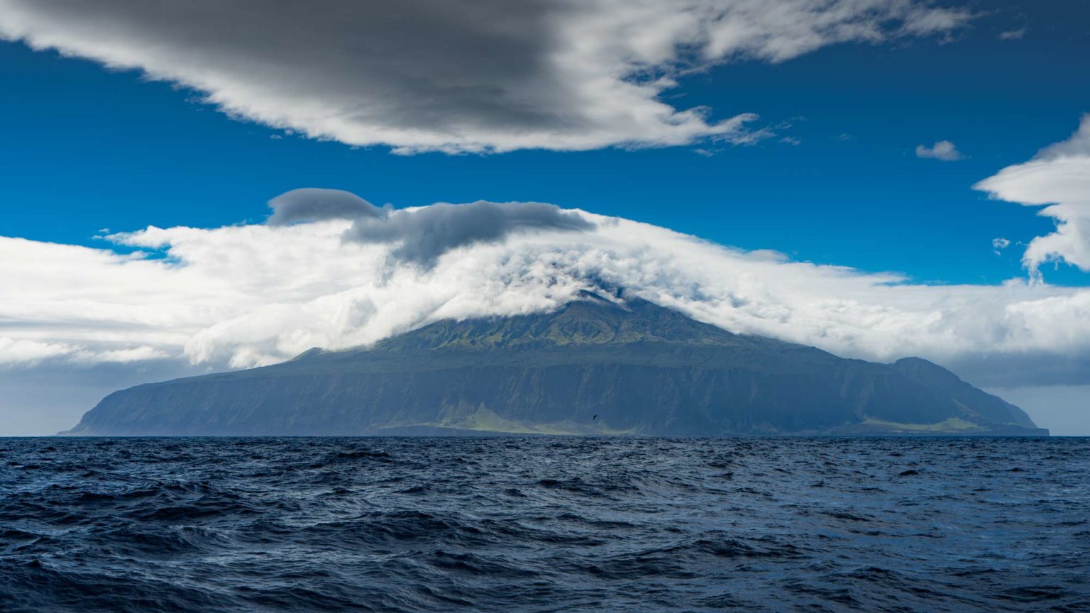 Moody analog film image of a large mountainous island rising above dark ocean waters, low clouds rolling over the peaks, blue-grey atmosphere throughout.